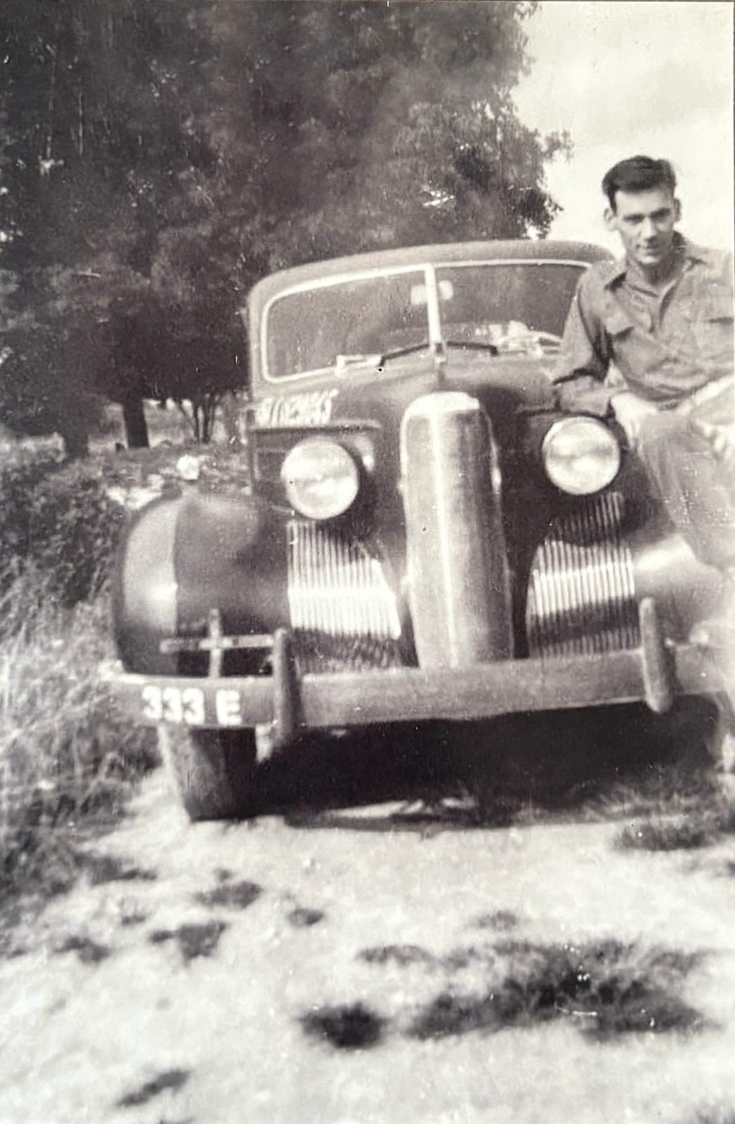 George perched on the fender of a requisitioned 1930s sedan, bumper marked '333 E.' Summer, leafy — somewhere in France.