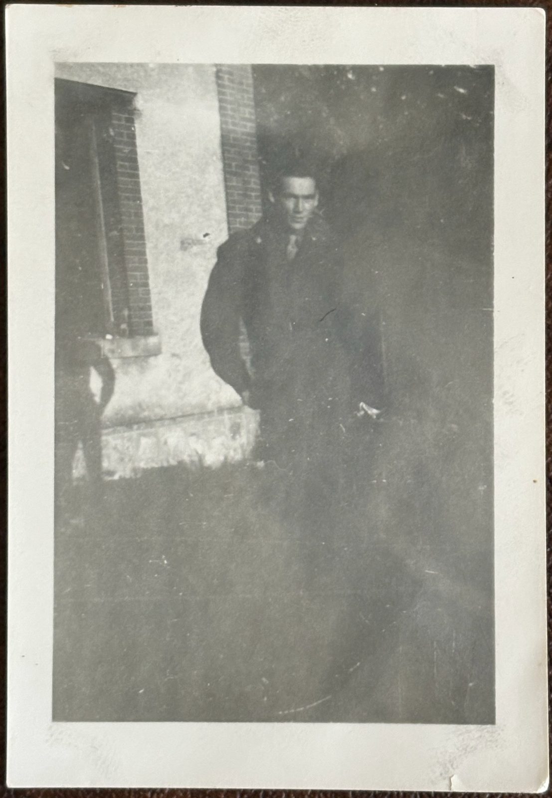 A soldier in a heavy overcoat against a brick corner in falling snow — the long wait overseas before D-Day.